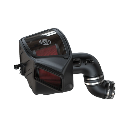S&B Filters cold air intake kit with a matte black enclosed airbox, a clear plastic inspection window on top showing a red pleated air filter inside, a secondary red-filtered front inlet, and a curved black intake tube fitted with polished metal hose clamps.