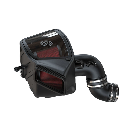 S&B Filters cold air intake kit with a matte black enclosed airbox, a clear plastic inspection window on top showing a red pleated air filter inside, a secondary red-filtered front inlet, and a curved black intake tube fitted with polished metal hose clamps.